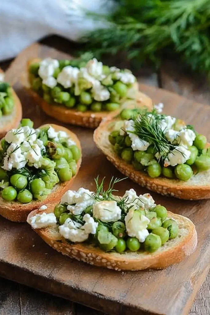 Knusprige Crostini mit grünen Erbsen, zerbröseltem Feta und frischem Dill auf einem rustikalen Holzbrett.
