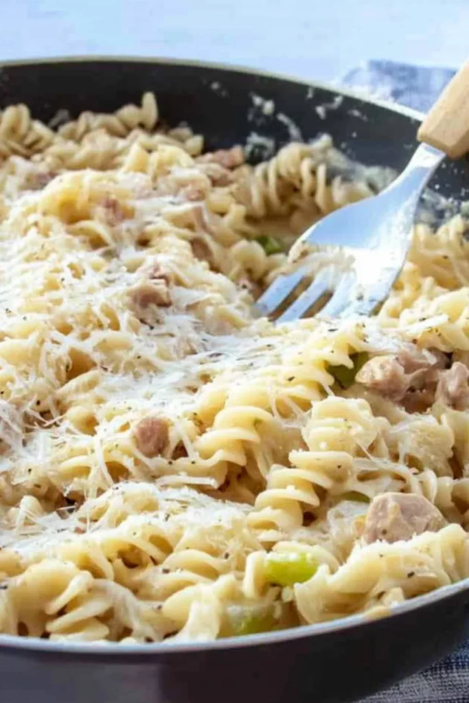 Pfanne mit Spiralnudeln in einer cremigen Crème-Fraîche-Sauce, garniert mit geriebenem Käse und kleinen Speck- oder Fleischstücken, mit einer Gabel, die in die Pasta getaucht ist.